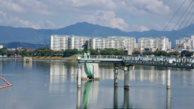 Soyang River Skywalk stretches over Uiam Lake with apartments and mountain ridges reflected in the calm water.