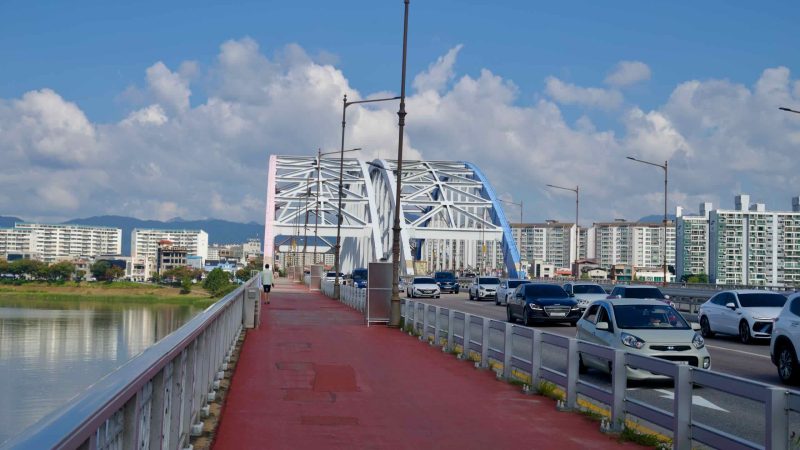 Pedestrians use the walkway beside traffic on Chuncheon’s arched bridge, with pastel-painted steel truss arches.