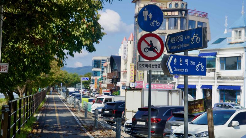 Direction signs for the Bukhangang Cycleway and Chuncheon Station stand beside a shaded riverside bike lane.