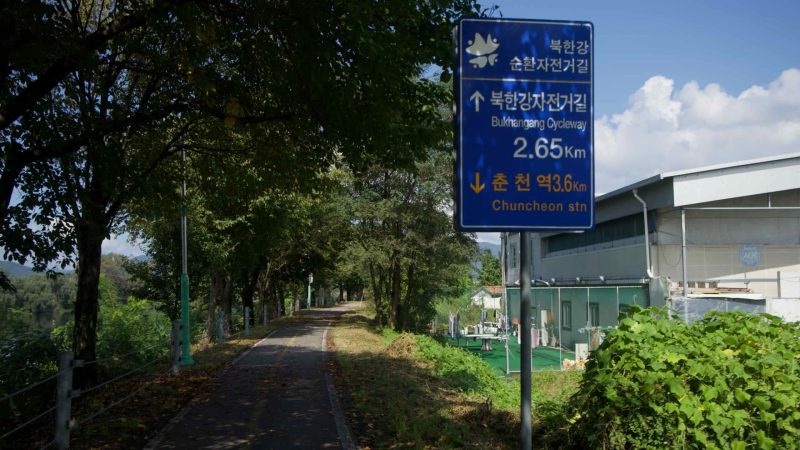 A blue route marker shows distances for the Bukhangang Cycleway and Chuncheon Station along a shaded levee.