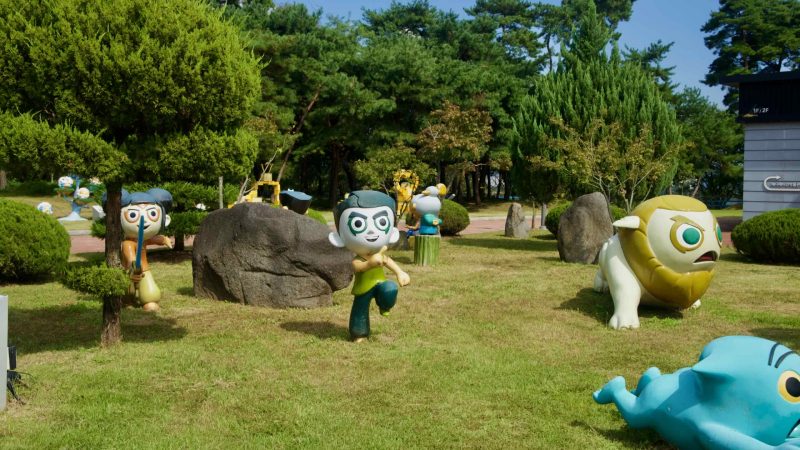 Wide view of cartoon figures around boulders and pines in the museum garden.