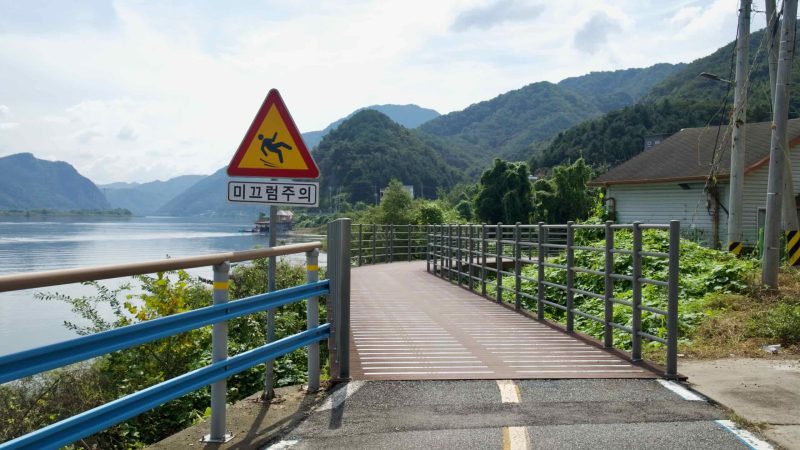 A slippery-surface warning sign stands by a boardwalk ramp on the river bike path in Chuncheon.