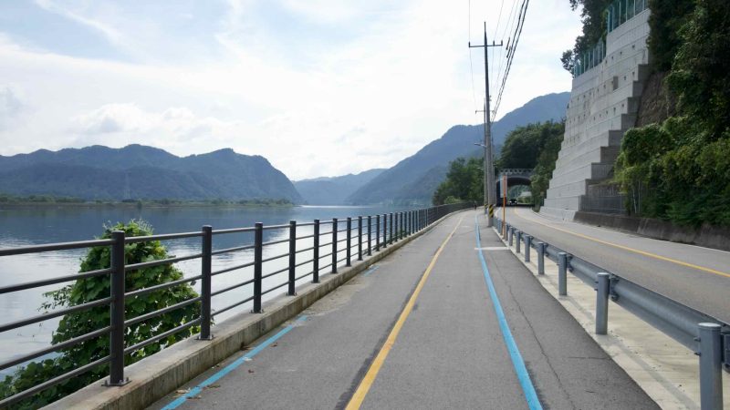 River bikeway beside a tiered retaining wall with a road tunnel ahead in Chuncheon.