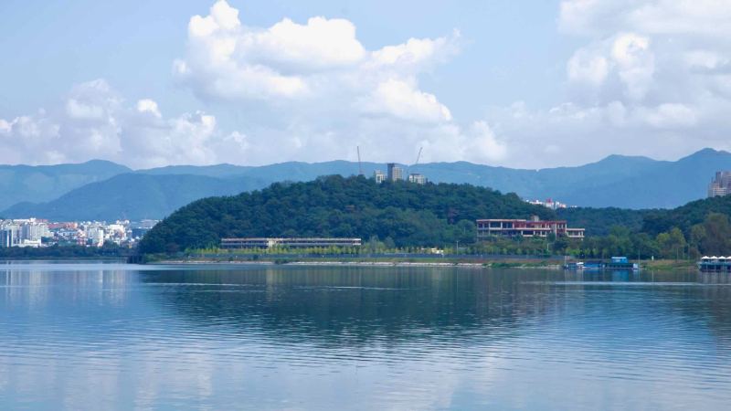 Across Uiam Lake, a wooded hill on Jungdo shows cranes and buildings above the lakeshore park.