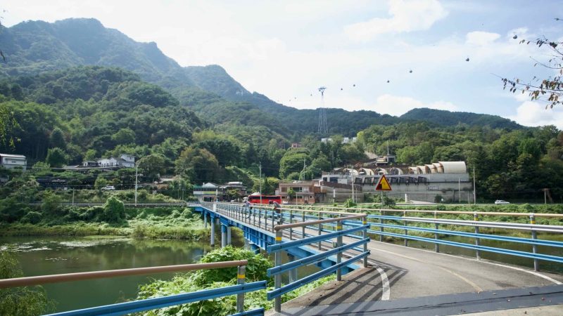 A blue bridge carries the bikeway across an inlet toward a hillside village.