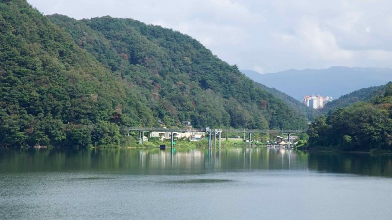 A narrow reach of Uiam Lake winds between forested slopes as distant apartments rise at the mouth of the valley.