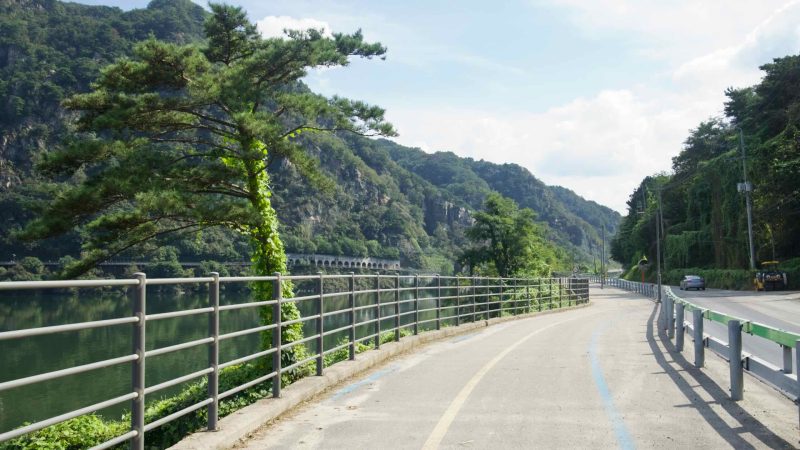 A lakeside bike path curves along Uiam Lake toward an arched gallery beneath the forested cliffs near Samaksan.