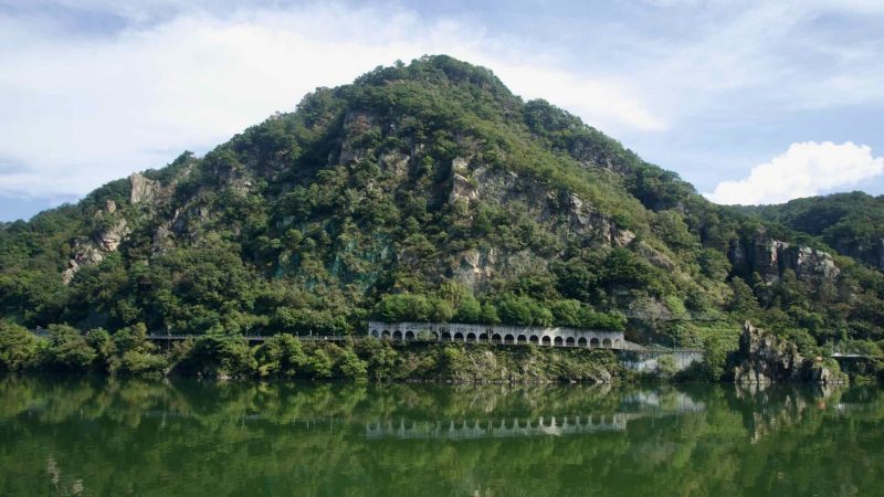 Arched cliff galleries trace the base of Samaksan above green Uiam Lake, their reflections repeating in the still water.