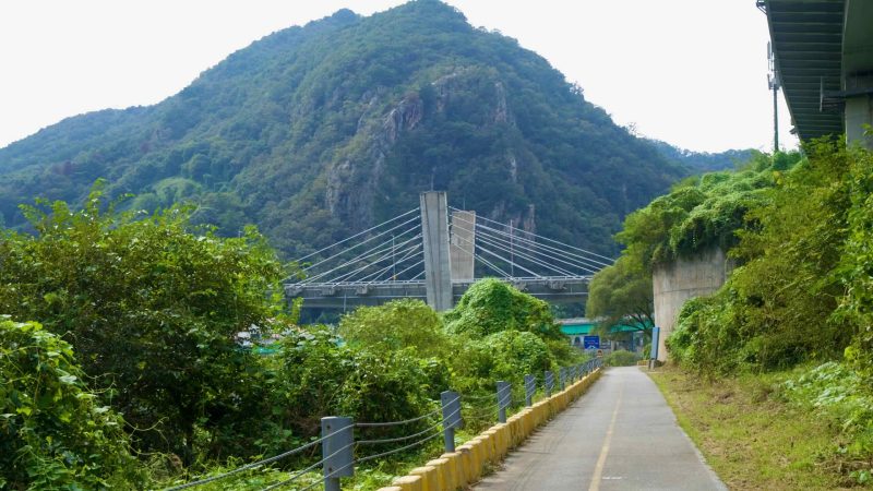 A riverside bike path curves toward Gangchon Grand Bridge beneath forested hills.