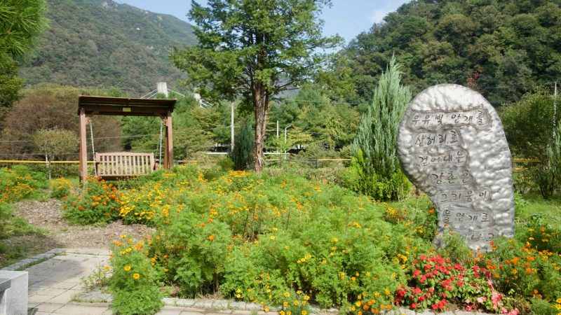 A small garden at Gangchon holds a swing bench amid marigolds and conifers beside a metallic poem stone marker.