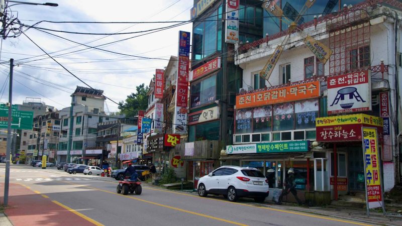 Dense signboards, cafés, and pensions crowd Gangchon’s main street along Gyeongchun Road as an ATV rolls past restaurants.