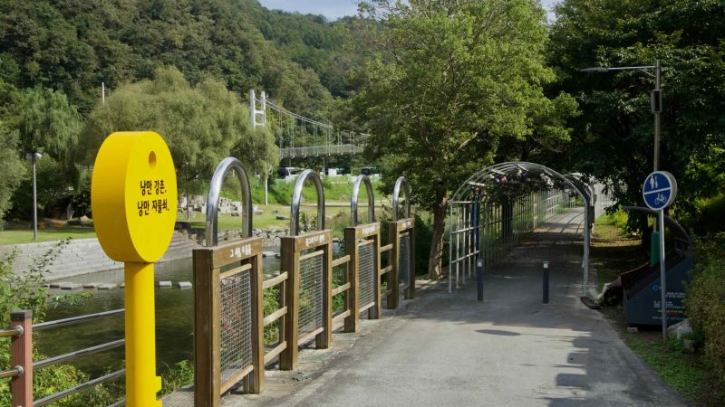 A yellow key sculpture marks a riverside promenade at Gangchon beside a metal lovelock tunnel and a suspension footbridge.