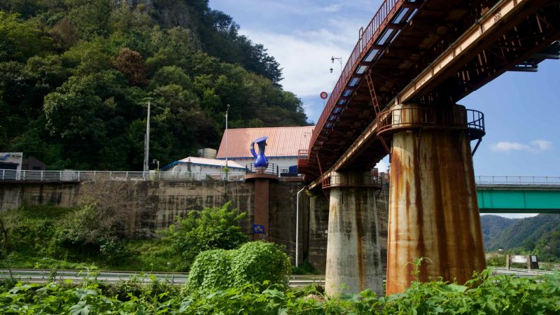 Rust-streaked piers of an old rail bridge stand above a tributary near Gangchon beside a riverside café and high-rises.