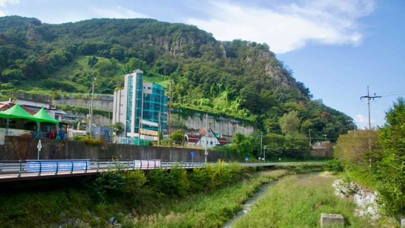 A narrow stream runs below a raised bikeway at Gangchon, backed by cliffs and vine-draped buildings and shacks.