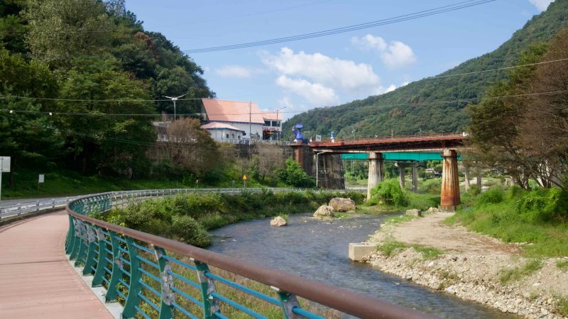 A curved riverside boardwalk follows a narrow stream toward an old reddish rail bridge near Gangchon’s green hills.