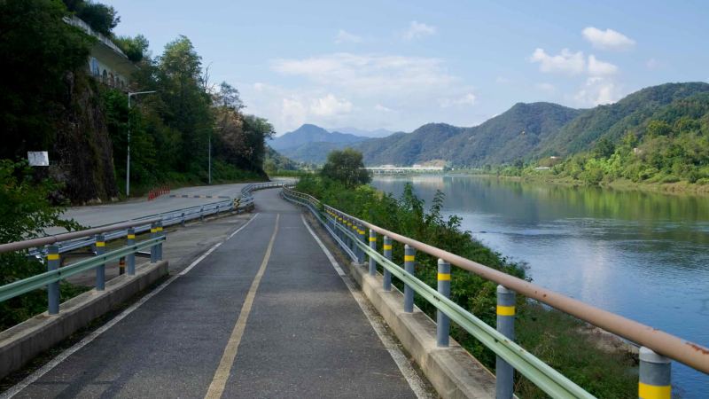 A protected bikeway bends along the Bukhangang beside guardrails and a quiet roadway with wooded hills beyond.