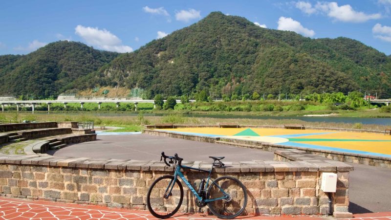 A road bike leans against a low stone wall at a colorful riverfront plaza in Gangchon below hills and a long viaduct.