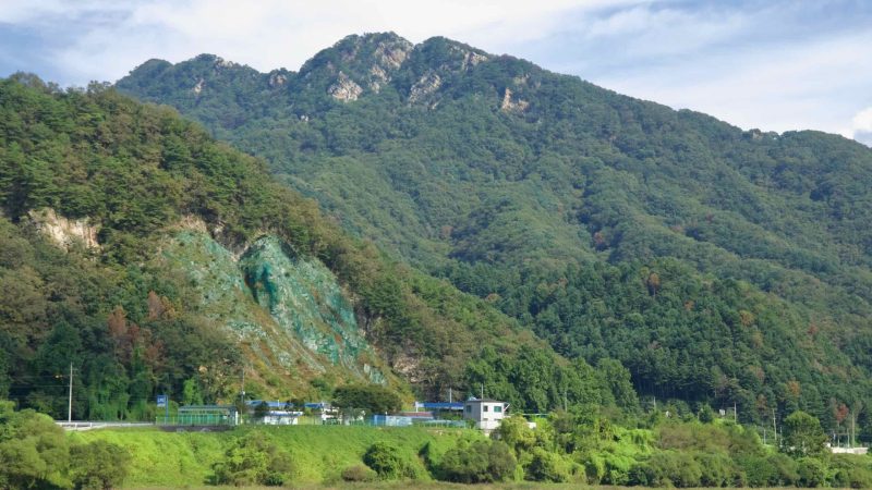 A rugged rock face with green-stained slopes rises above the riverside beneath thick forest and peaks near Gangchon.