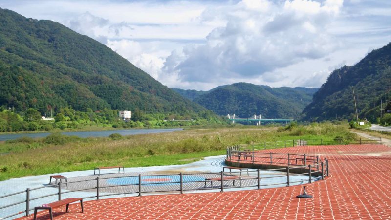 Benches and a patterned plaza overlook wetlands on the North Han River with Gangchon Grand Bridge in the distance.