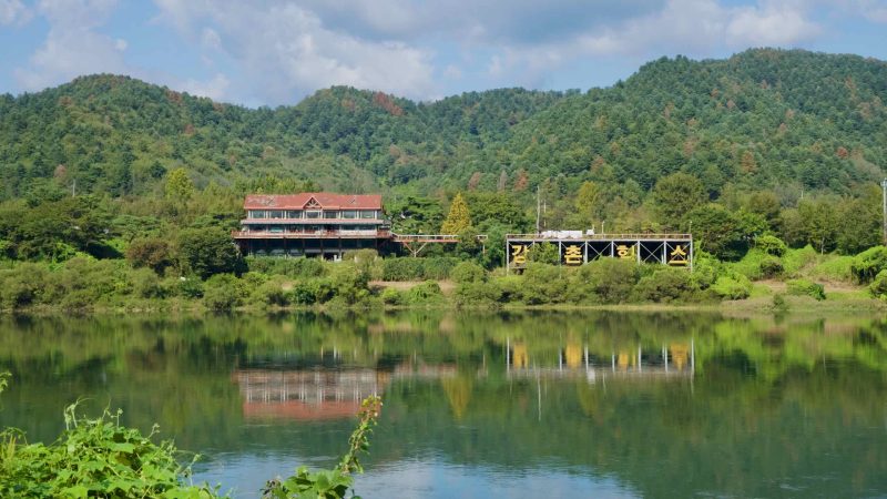 A riverside lodge and hillside sign reflect in the calm Bukhangang.