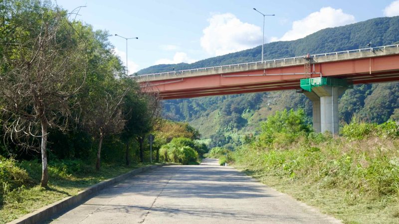 A quiet ramp passes under a red river bridge beside trimmed brush.
