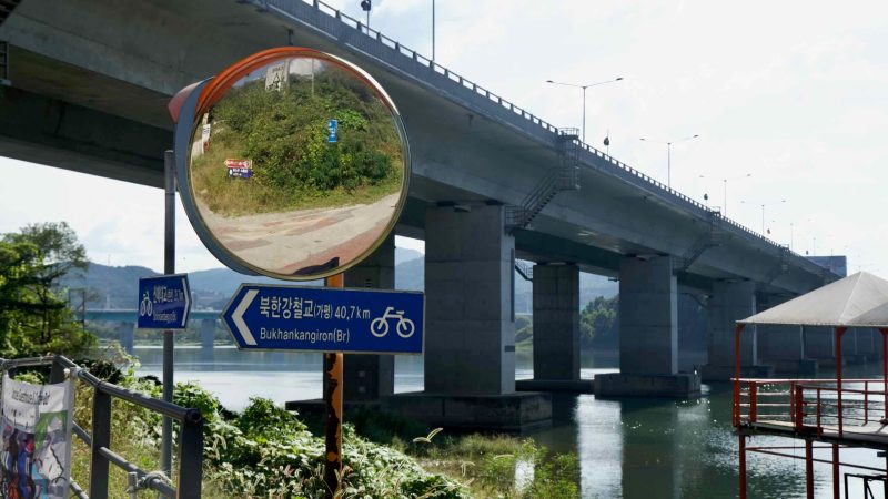 A convex mirror reflects the bikeway beneath a tall road bridge.