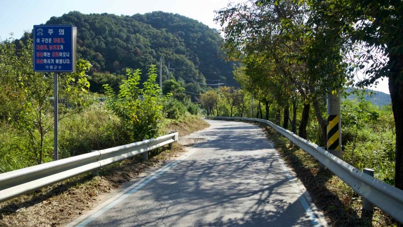 A narrow riverside lane of the Bukhangang Bike Path curves past a blue caution sign and roadside trees beside Daljeon Stream.