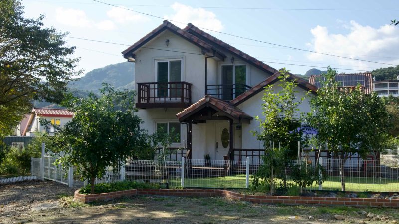 A two-story white house with tile roofs and a small garden stands behind a low fence.