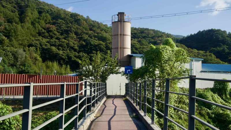A pedestrian walkway beside an industrial yard leads toward a tall storage silo.
