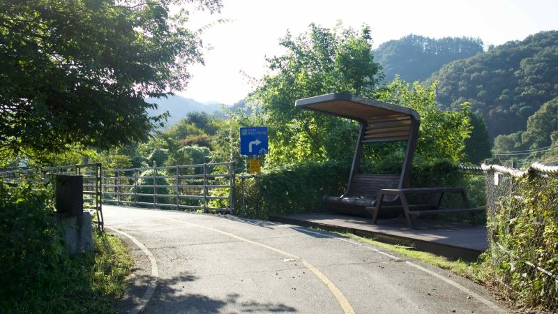 A wooden rest shelter and blue wayfinding sign stand beside a curving stretch of bikeway.