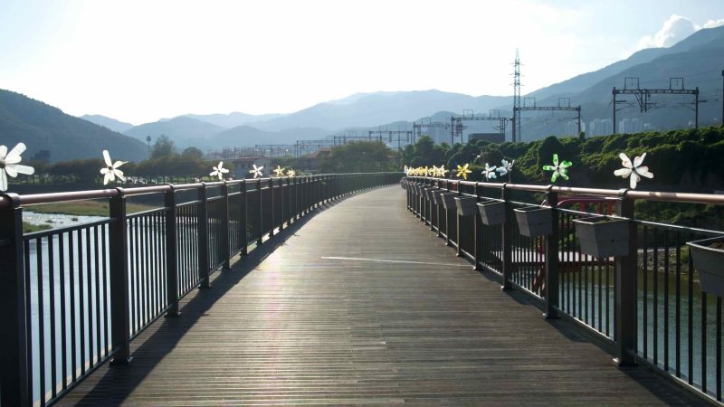 A riverside pedestrian bridge lined with decorative pinwheels crosses toward Cheongpyeong as mountains layer in hazy distance.