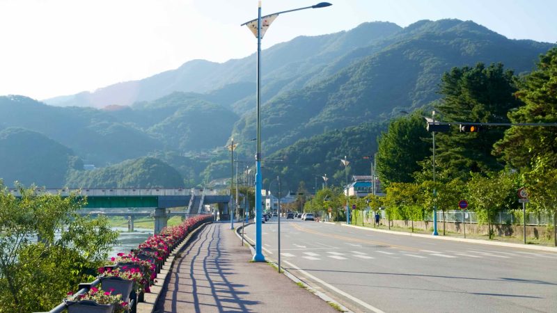 Planter boxes of bright flowers line a riverside promenade and roadway in Cheongpyeong, with a low bridge over the stream ahead.