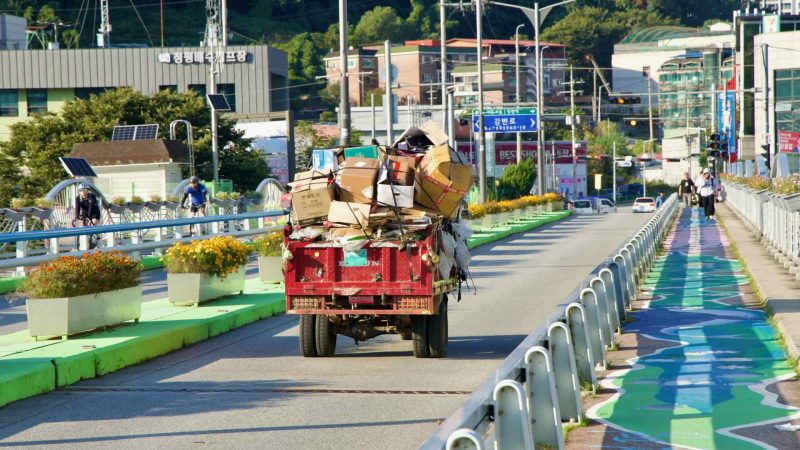 A recycler’s cart loaded with boxes moves along Cheongpyeong Bridge, as cyclists and pedestrians use the adjacent painted lane.
