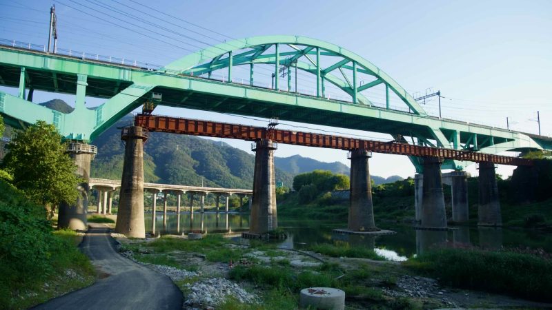 Under the aquamarine arch of the Gyeongchun Line bridge at Cheongpyeong, an older rust-colored rail span and distant hills align.