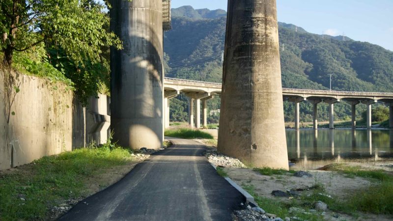 A fresh strip of asphalt threads between massive bridge piers on the riverside path below Cheongpyeong 1 Bridge, with mountains rising ahead.
