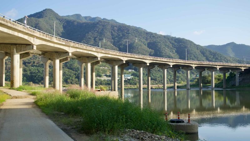 A sweeping concrete viaduct curves above calm water on the Bukhan River near Cheongpyeong, its repeating piers reflected below.