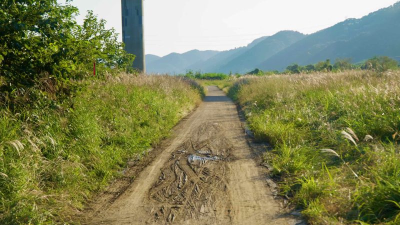 A rutted dirt trail climbs between thick grasses near a solitary bridge pillar, with hazy ridges stacking across the Bukhan River valley.