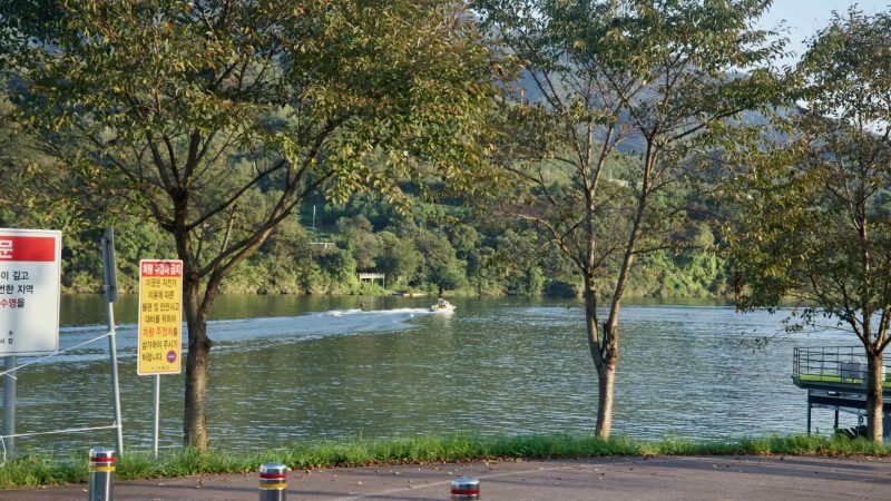 Signs and bollards mark a North Han River access point as a speedboat passes in the distance.