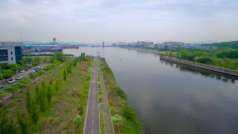 A straight stretch of the Ara Waterway runs west toward the sea lock, with bike and pedestrian paths lining the canal amid greenery and port infrastructure in the distance.