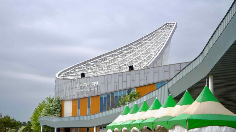 The Ara Incheon Passenger Terminal features a distinctive sail-shaped roof and bold architectural design, standing beside green-topped tents set up along the walkway.