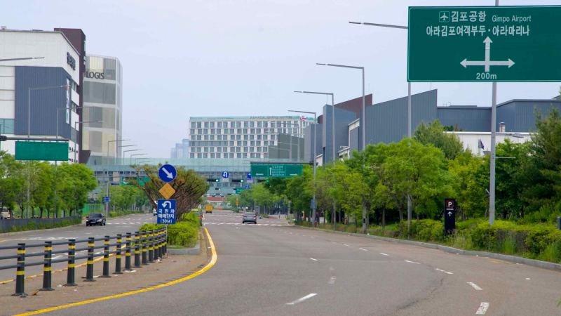 A wide boulevard leads toward the Ramada Gimpo Hotel, flanked by warehouses and signs directing travelers to Gimpo Airport, Ara Marina, and Ara Gimpo Passenger Terminal.