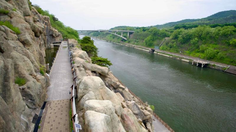 From midway up the rockface at Ara Falls, a walkway and dramatic view unfold over the Ara Canal, with bridges, tree-lined slopes, and riverside trails extending into the distance.