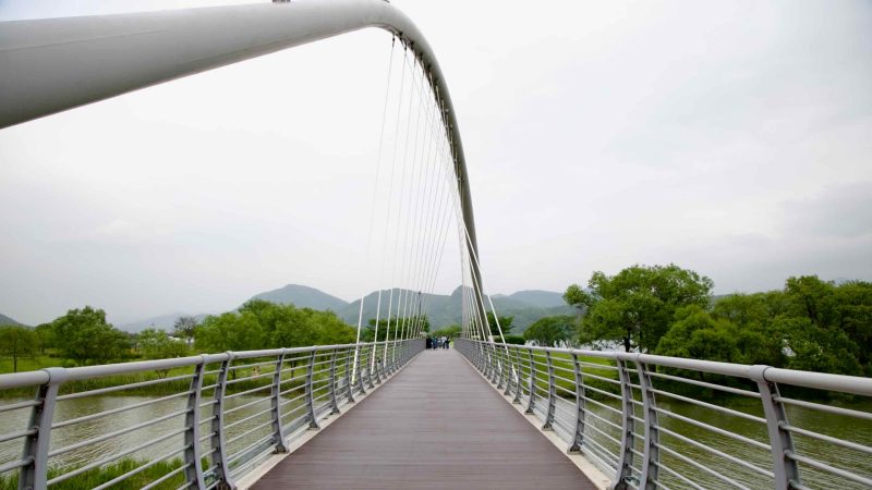 Baetnari Bridge, a curved pedestrian span, stretches over calm wetlands in Namyangju Water Garden, connecting scenic paths beneath forested mountains.