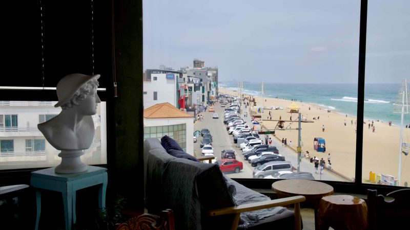 A cozy cafe interior with eclectic decor overlooks Gangneung Coffee Street and the beach, offering a scenic view of the road, sand, and ocean through large windows.