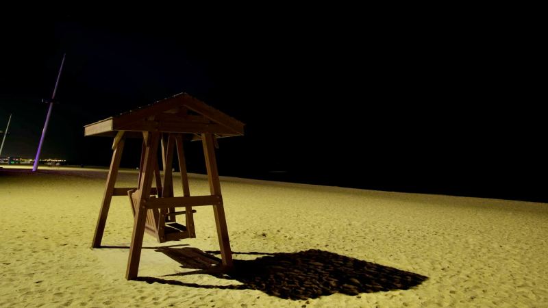 A wooden swing sits on the sandy shores of Gyeongpodae Beach under the night sky, casting a shadow in the dim light, evoking a calm and tranquil coastal scene.