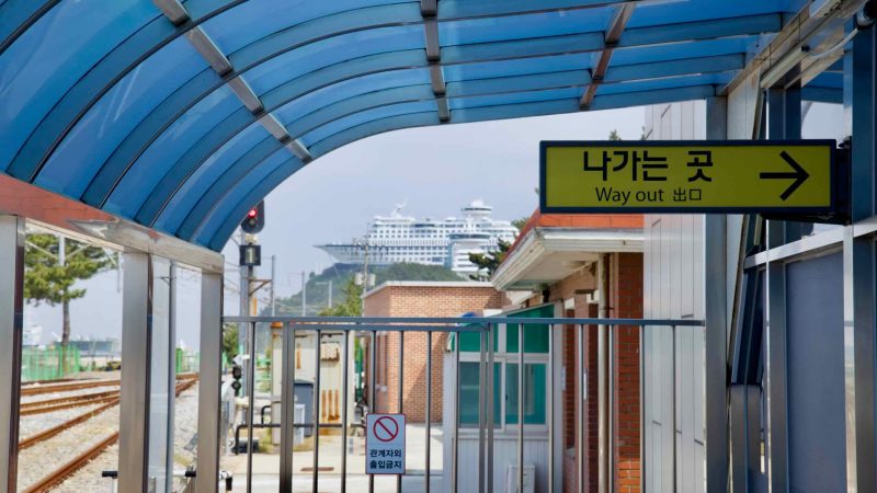 Yellow "Way Out" sign under the blue canopy at Jeongdongjin Station, with the Sun Cruise Resort visible.