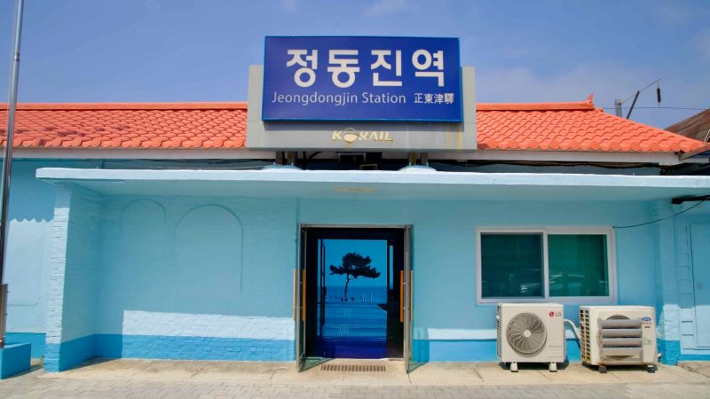 Jeongdongjin Station's pastel blue facade with a red roof and signs in Korean and English.