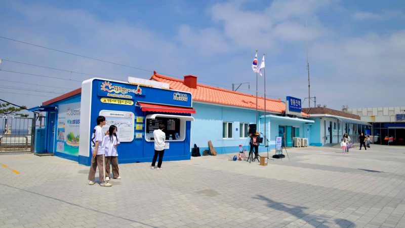 Visitors outside the sky-blue Jeongdongjin Station building with an orange roof and national flags.