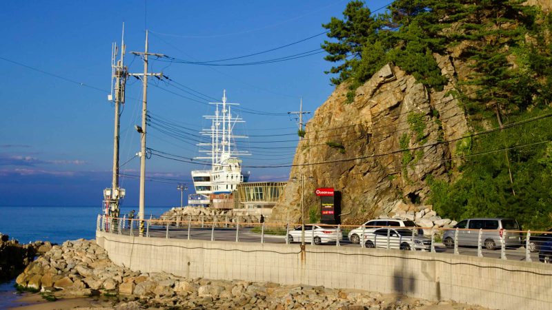 Along the coastal road near Jeongdongjin, a unique building in the shape of a ship with fake masts overlooks the rocky coast and waters below, housing a fish restaurant and coffee shop.