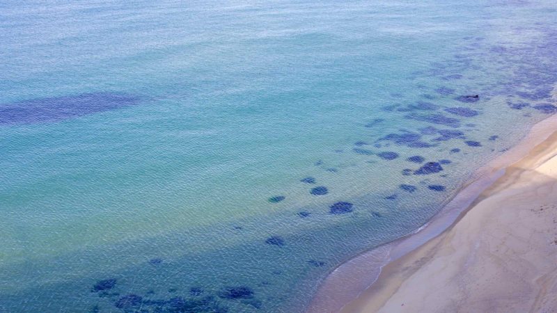 Aerial view of Mangsang Beach reveals the transparent East Sea waters meeting the sandy beach, with visible sand patterns and seaweed beneath the gentle waves.